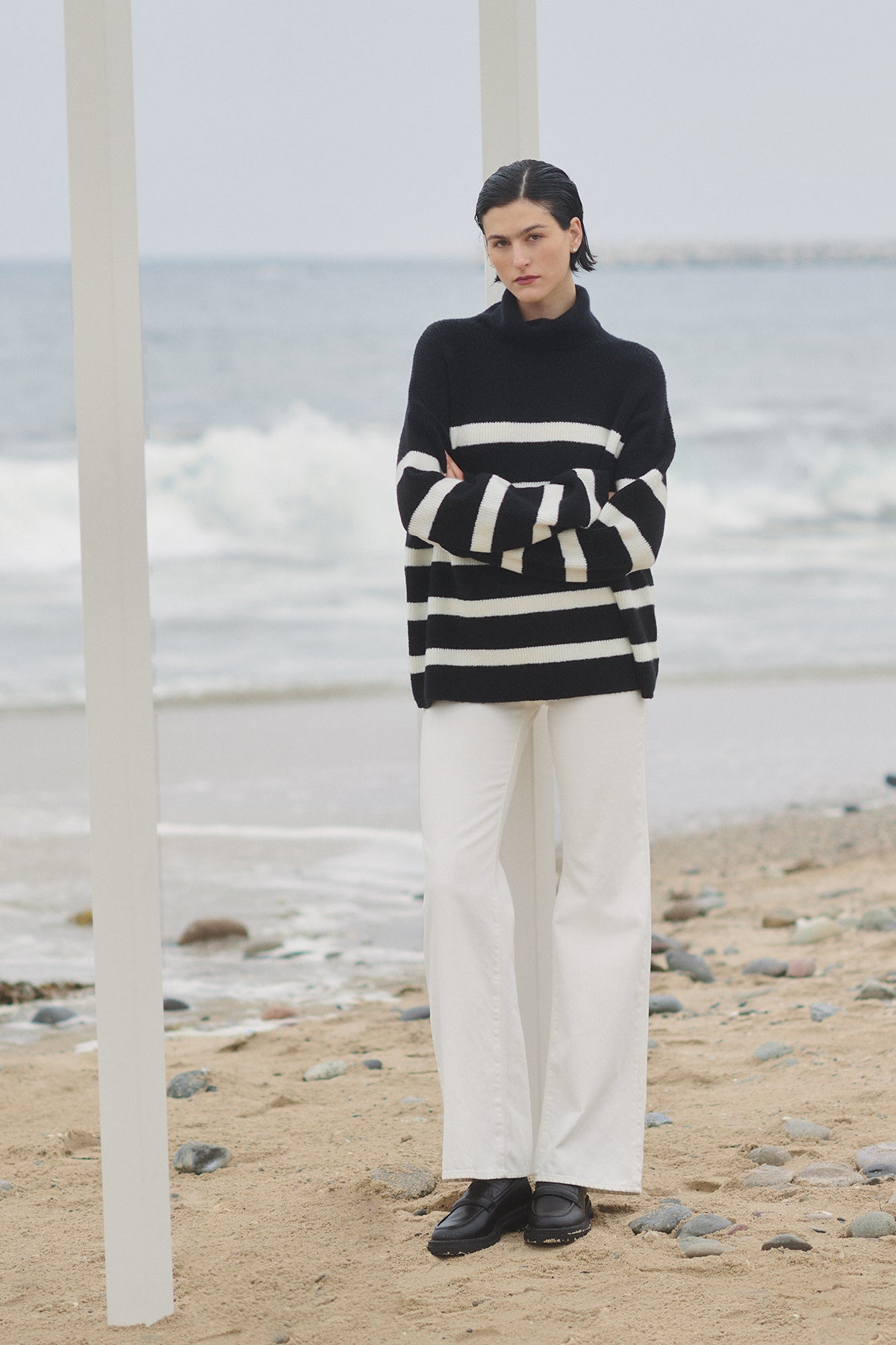   A woman wearing an oversized fit, Velvet by Jenny Graham's ENCINO SWEATER, a black and white striped turtleneck sweater, and white pants on the beach. 