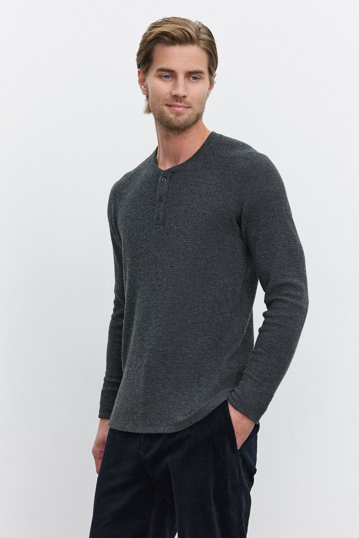 A man with light brown hair stands against a plain white background, wearing the AMBROSE HENLEY by Velvet by Graham & Spencer. The long-sleeve henley shirt in dark gray is made from cozy thermal fabric and paired with dark pants. The three-button placket adds a touch of style as he has his hands in his pockets.-37893849284801