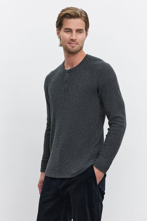 A man with light brown hair stands against a plain white background, wearing the AMBROSE HENLEY by Velvet by Graham & Spencer. The long-sleeve henley shirt in dark gray is made from cozy thermal fabric and paired with dark pants. The three-button placket adds a touch of style as he has his hands in his pockets.