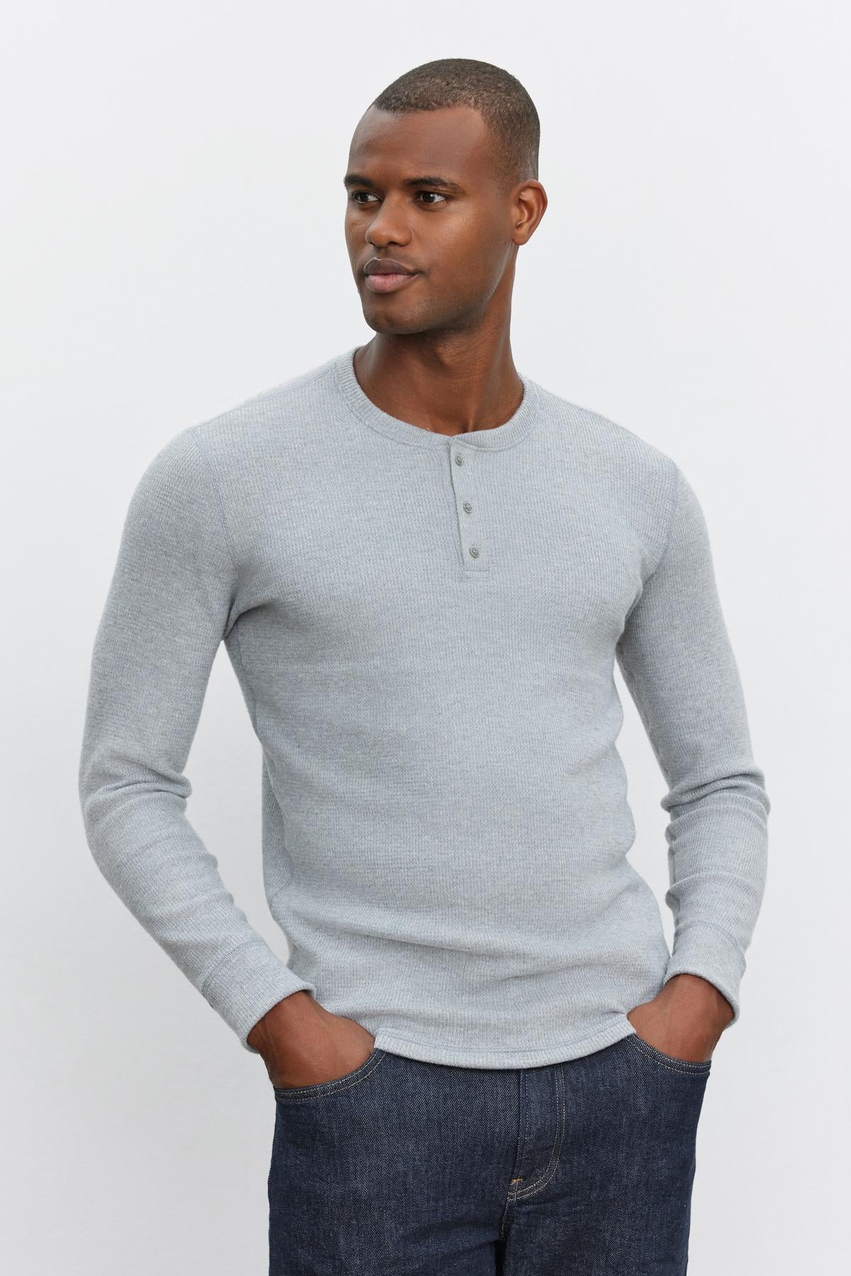   A man wears the AMBROSE HENLEY, a light grey long-sleeve shirt by Velvet by Graham & Spencer, and dark blue jeans, standing with hands in his pockets against a plain white background. 