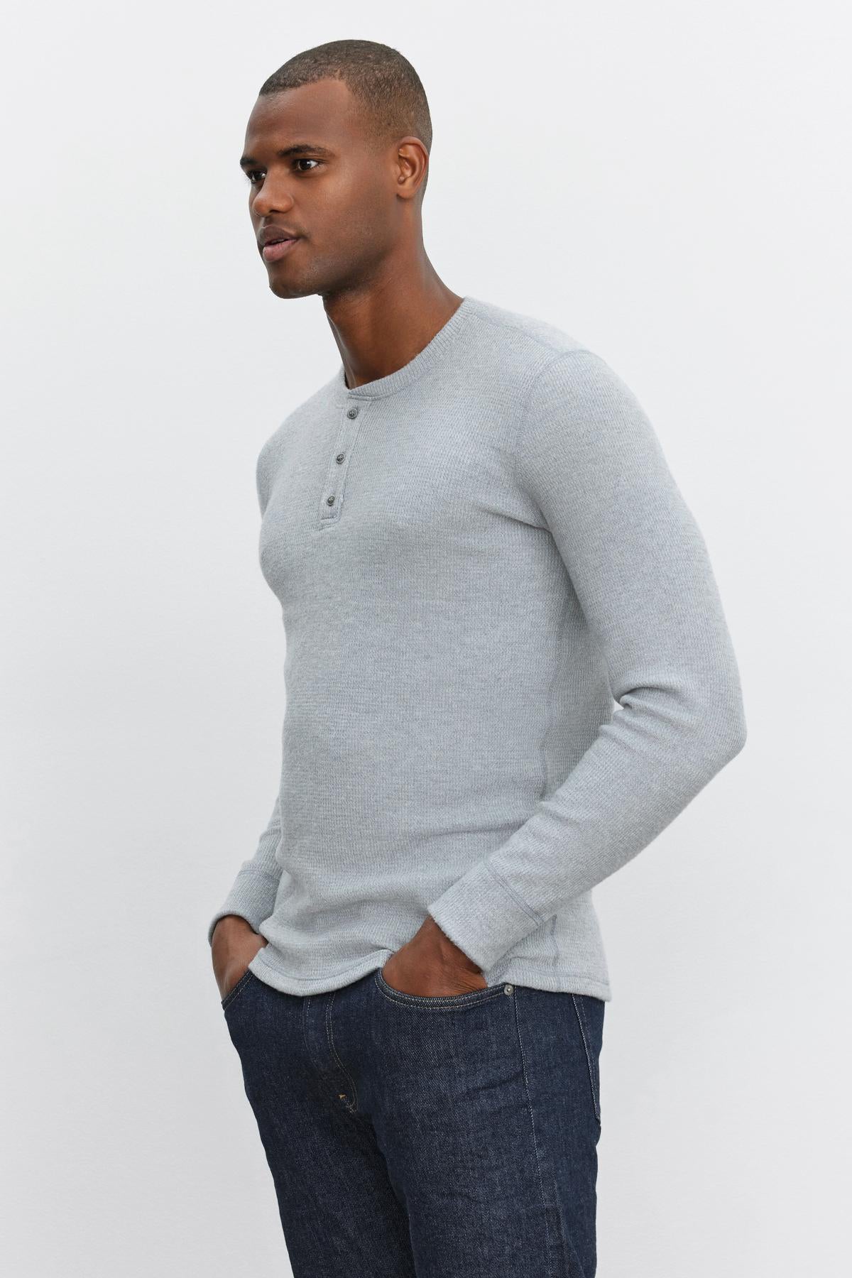   A man in a light grey AMBROSE HENLEY by Velvet by Graham & Spencer, paired with blue jeans, stands against a plain white background, his hands in his pockets as he looks to the side. 