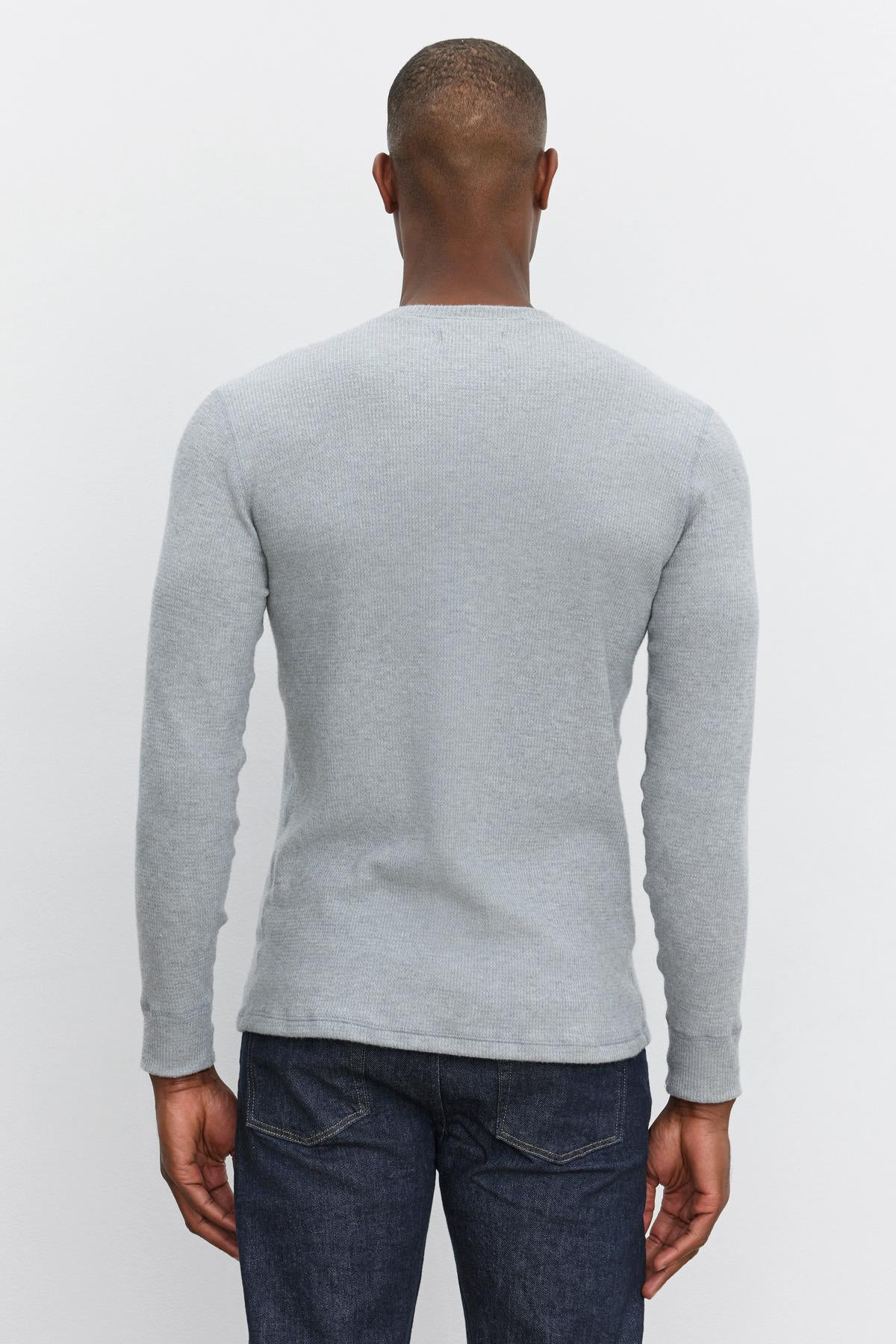   A person is standing with their back to the camera, wearing a light grey AMBROSE HENLEY by Velvet by Graham & Spencer and dark blue jeans. 