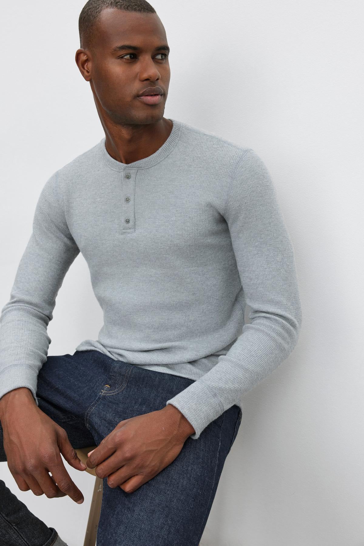   A man in the AMBROSE HENLEY by Velvet by Graham & Spencer and dark jeans sits on a wooden stool against a plain white background. 