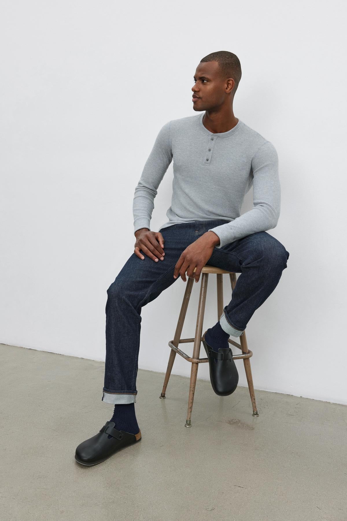   A person wearing an AMBROSE HENLEY by Velvet by Graham & Spencer, made of triblend cozy thermal fabric in light gray, paired with dark jeans and black clogs, sits on a wooden stool against a white background. 