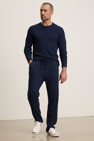 A man stands against a plain background wearing Velvet by Graham & Spencer's OZZY SWEATPANT—relaxed fit joggers with a drawstring waist—paired with a navy blue long-sleeve sweater and white sneakers.