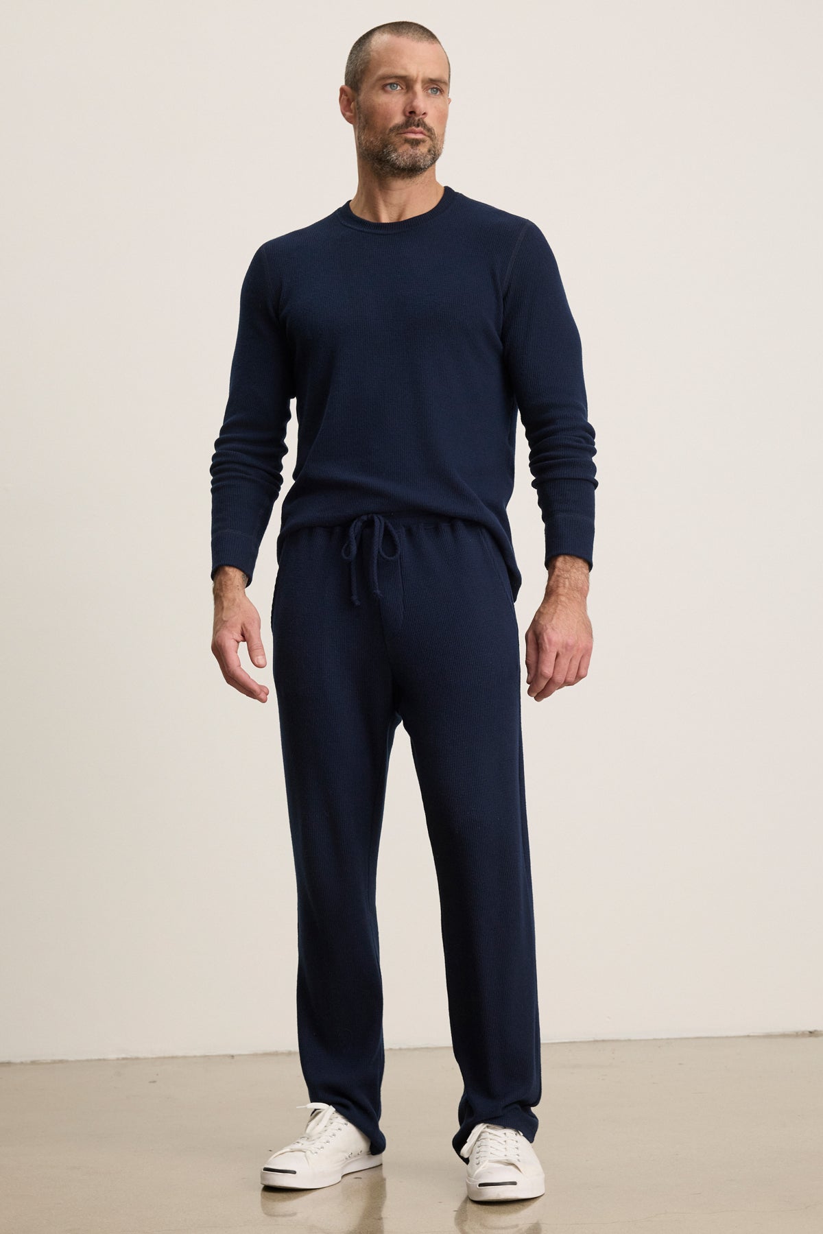 A man stands indoors against a plain light background wearing Velvet by Graham & Spencer's OZZY SWEATPANT in navy, paired with a relaxed fit long-sleeve sweater and white sneakers.