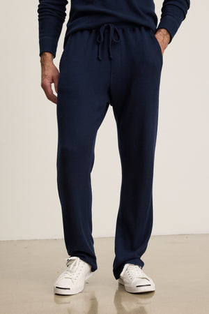 A person wearing Velvet by Graham & Spencer's OZZY SWEATPANT in navy blue with white sneakers stands on a light-colored floor against a plain background.