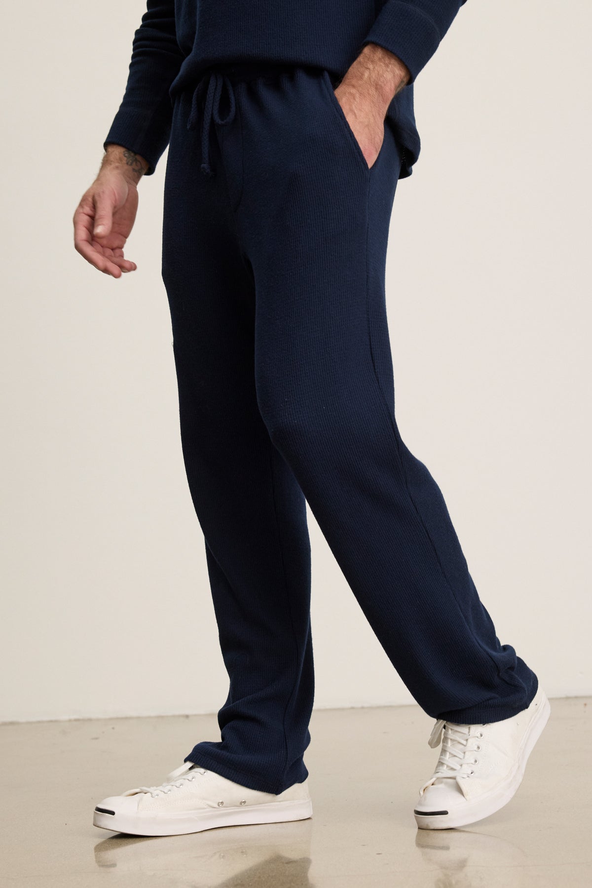 A person wearing Velvet by Graham & Spencer's OZZY SWEATPANT in navy blue with a drawstring waist and white sneakers stands on a light-colored floor, one hand casually in their pocket.-39957109735617