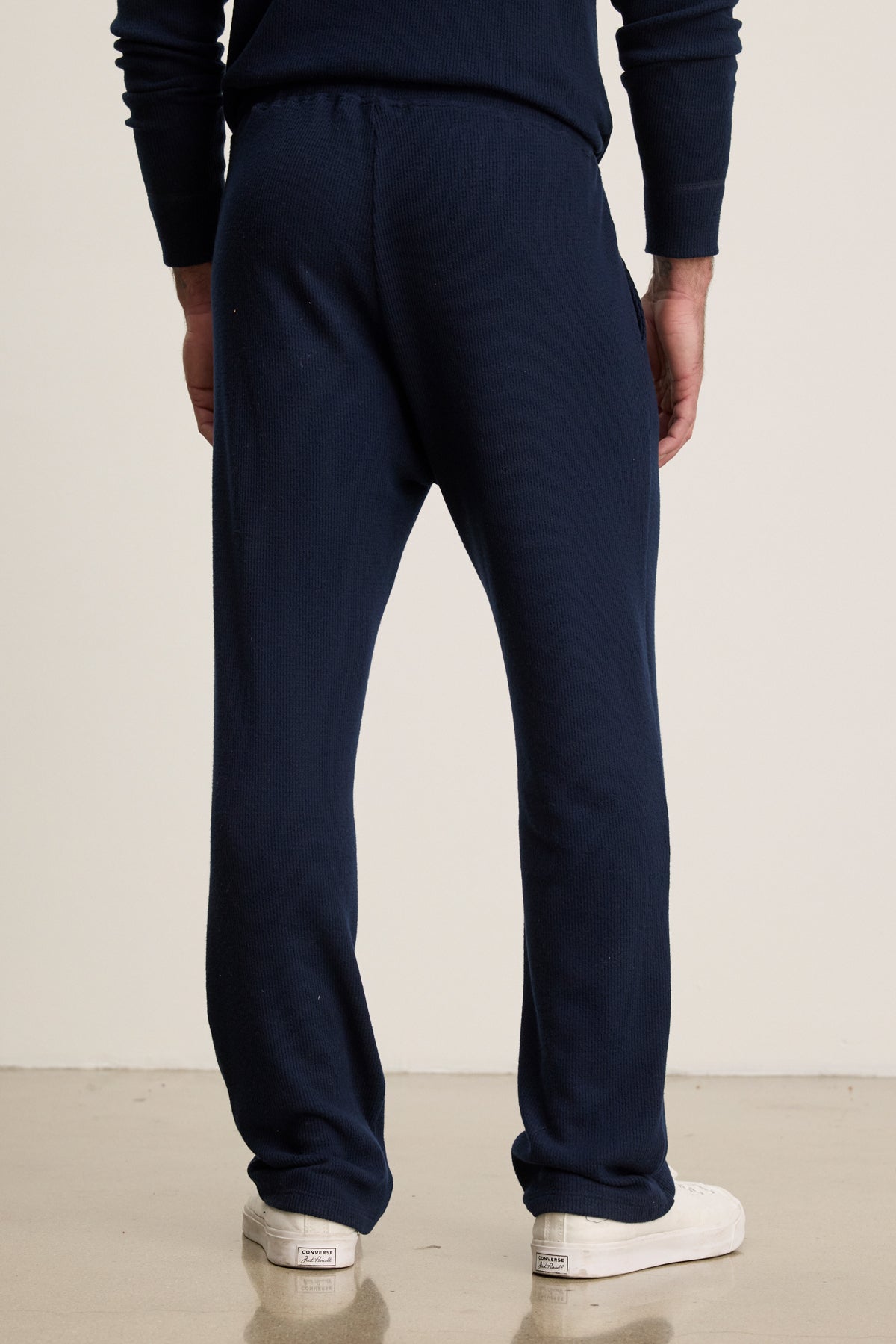 Someone stands on a light-colored floor facing away from the camera, wearing Velvet by Graham & Spencer's OZZY SWEATPANT in relaxed fit navy blue with a drawstring waist, paired with white sneakers.-39957109768385