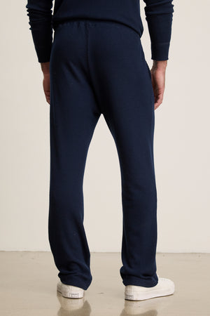 Someone stands on a light-colored floor facing away from the camera, wearing Velvet by Graham & Spencer's OZZY SWEATPANT in relaxed fit navy blue with a drawstring waist, paired with white sneakers.