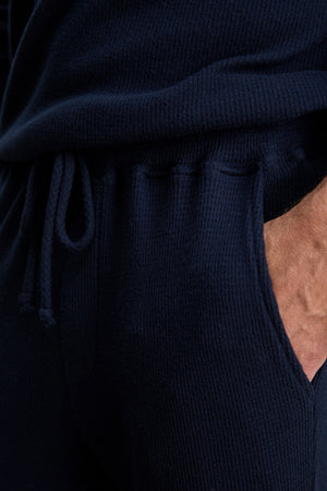 A person wearing a blue sweater and the OZZY SWEATPANT by Velvet by Graham & Spencer, featuring a relaxed fit and drawstring waist, with his hand in his pocket.