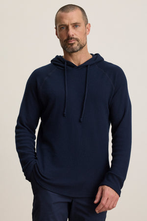 A man with short hair and a beard models the ROMEO HOODIE by Velvet by Graham & Spencer in navy blue, relaxed fit, paired with dark pants, standing against a plain light background.