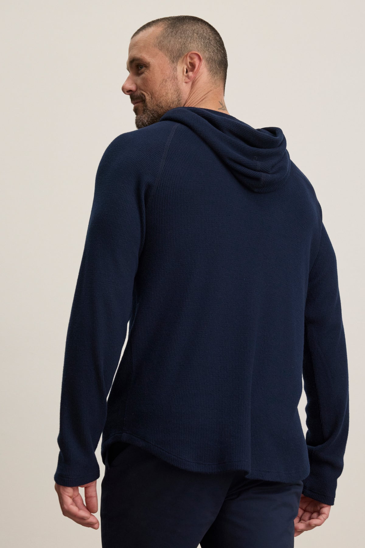 A man wears the ROMEO HOODIE by Velvet by Graham & Spencer—a navy blue, relaxed-fit pullover—as he stands facing away and looks over his left shoulder against a plain background.-39957110685889