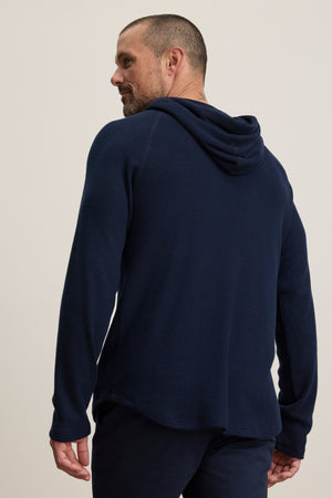 A man wears the ROMEO HOODIE by Velvet by Graham & Spencer—a navy blue, relaxed-fit pullover—as he stands facing away and looks over his left shoulder against a plain background.