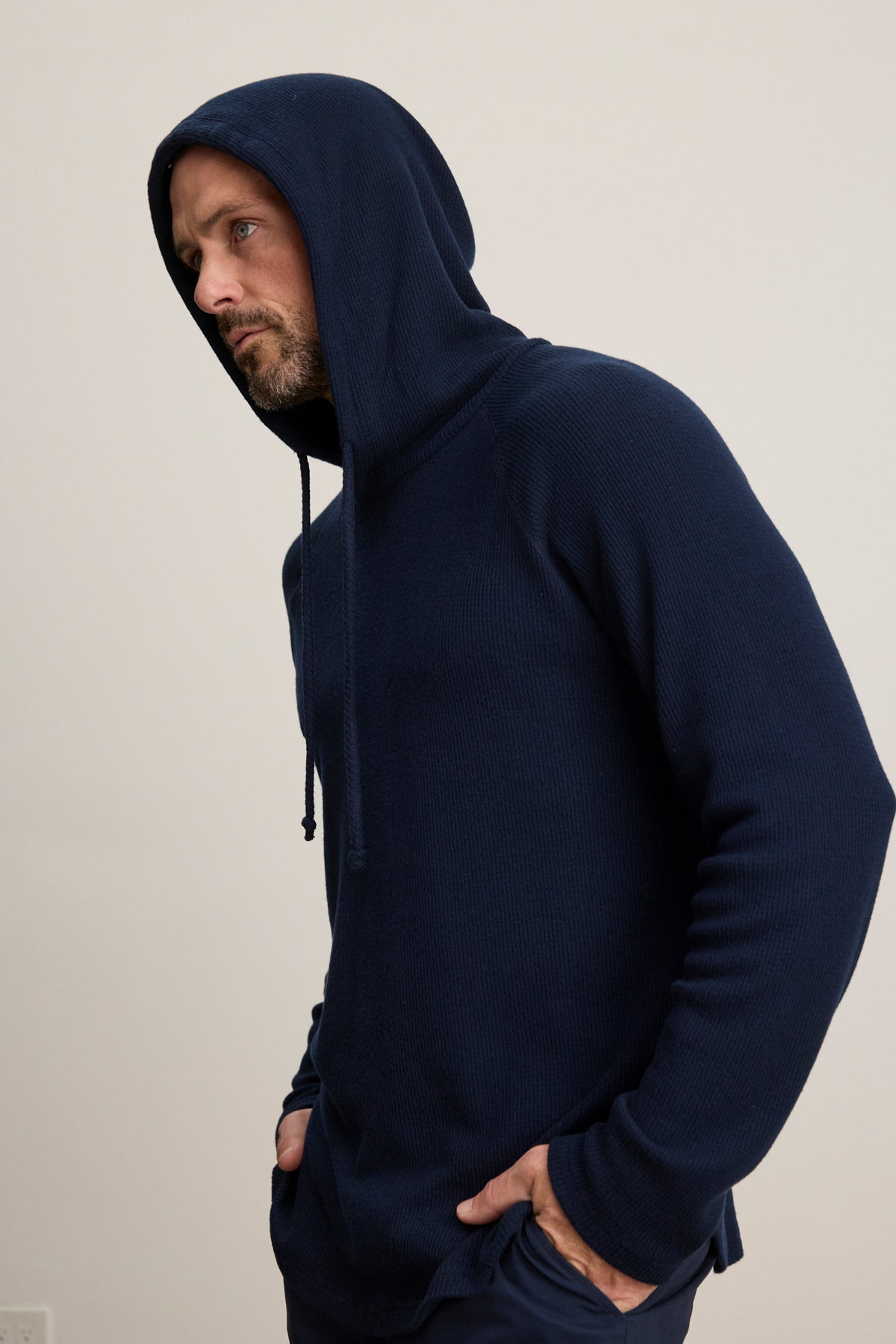 A man models the ROMEO HOODIE by Velvet by Graham & Spencer in navy blue, standing with hands in his pockets against a plain light background and highlighting its relaxed fit.-39957110784193
