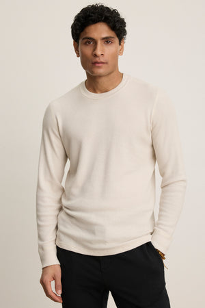 A man stands against a plain background wearing the TOBIAS CREW, a long-sleeve off-white textured shirt with a classic neckline from Velvet by Graham & Spencer, paired with dark pants, looking directly at the camera.