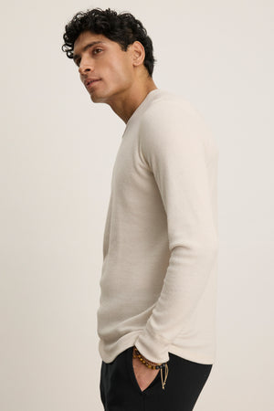 A man with short curly hair wears the TOBIAS CREW by Velvet by Graham & Spencer, a cozy beige triblend thermal long-sleeve shirt, paired with black pants, standing in profile against a plain light background.