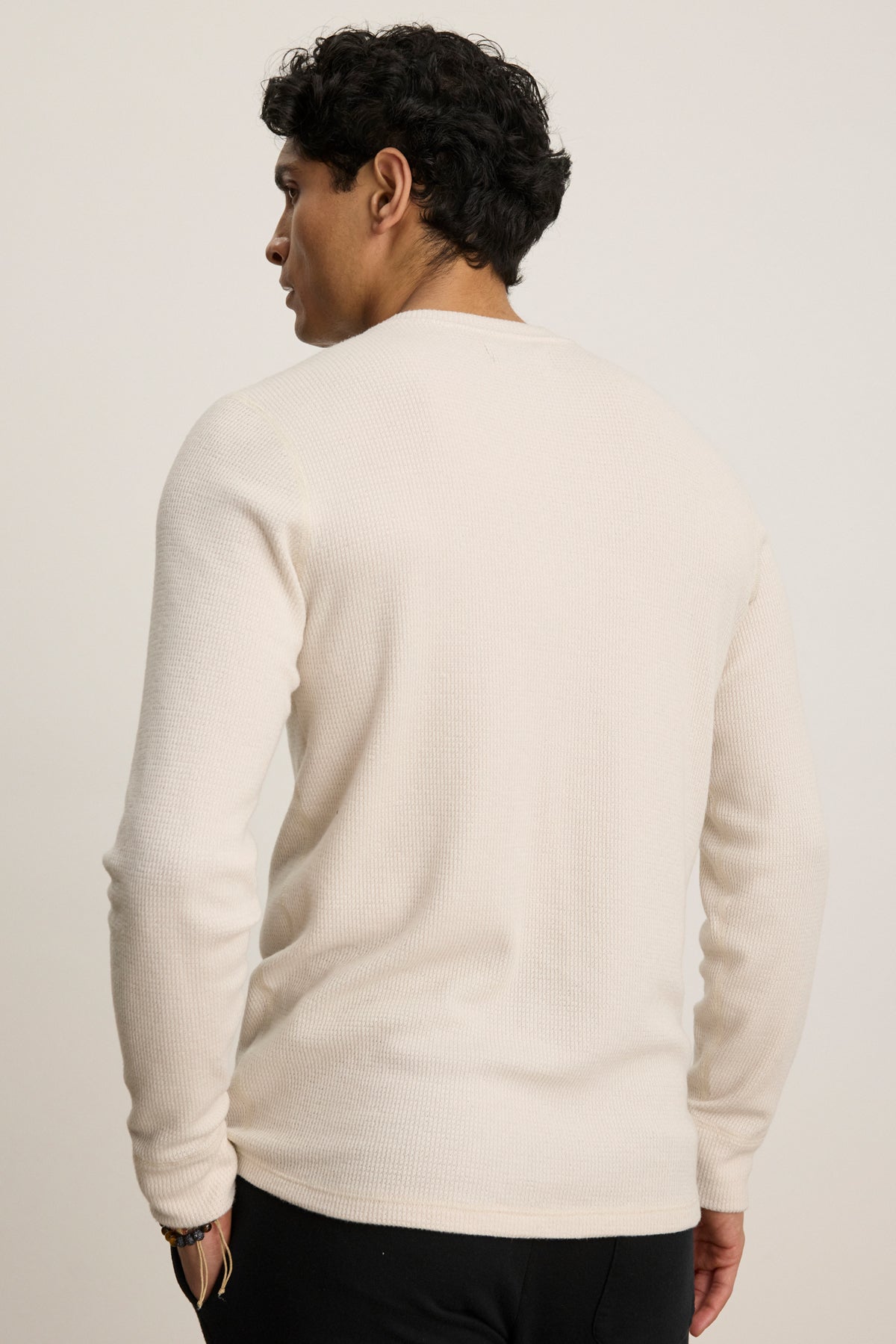 A man with dark hair is shown from the back wearing the Velvet by Graham & Spencer TOBIAS CREW, a long-sleeve off-white sweater with a classic neckline, paired with black pants against a plain light background.-39543546314945