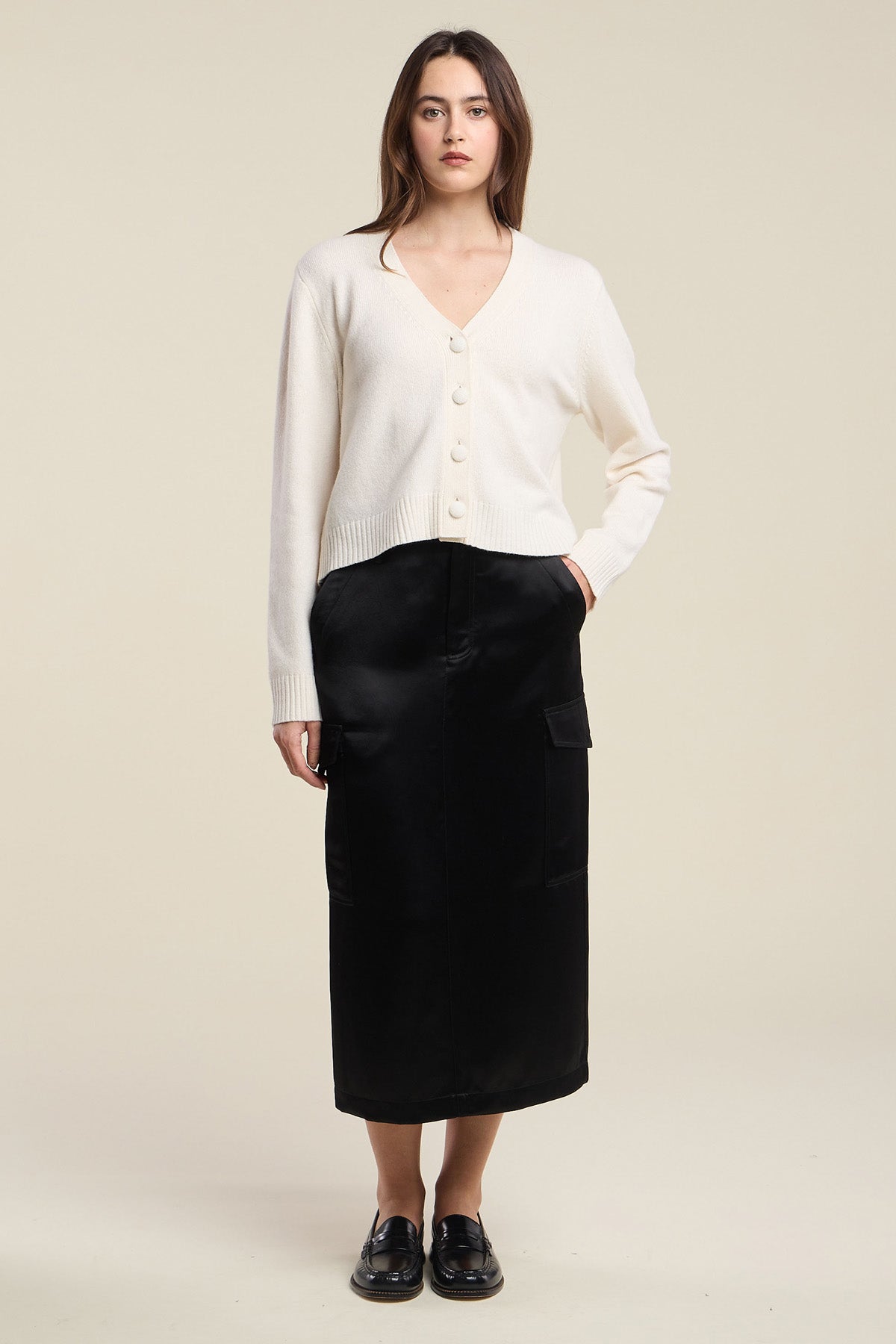 A woman stands against a plain background wearing the ETNA CARDIGAN by Velvet by Graham & Spencer—a white wool blend cardigan featuring a V-neckline and tailored fit—paired with a long black skirt with pockets and black loafers.-42063738208449