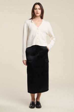 A woman stands against a plain background wearing the ETNA CARDIGAN by Velvet by Graham & Spencer—a white wool blend cardigan featuring a V-neckline and tailored fit—paired with a long black skirt with pockets and black loafers.