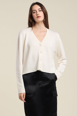 A woman wears the ETNA CARDIGAN by Velvet by Graham & Spencer—a white, wool blend cardigan featuring a V-neckline and tailored fit—paired with a black skirt, standing with one hand in her pocket against a plain background.