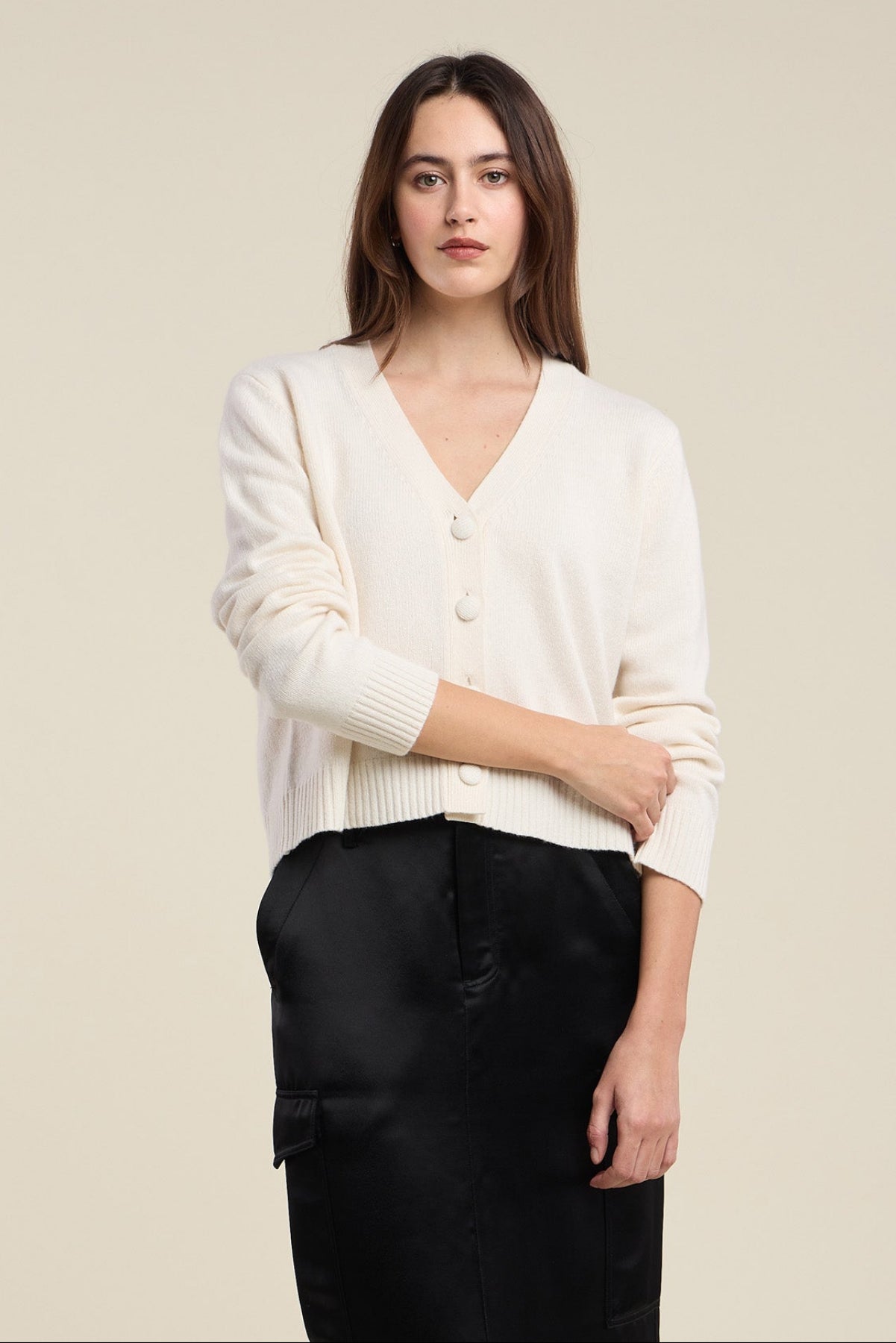 A woman with long brown hair wears the ETNA CARDIGAN by Velvet by Graham & Spencer—a cream wool blend cardigan featuring large buttons and a V-neckline—paired with a black skirt, standing against a beige background.
