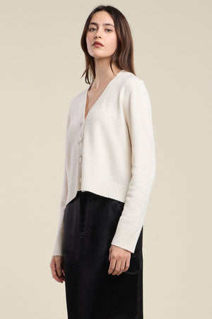 A person wearing the ETNA CARDIGAN by Velvet by Graham & Spencer—a cream-colored wool blend piece featuring a tailored fit and V-neckline—is paired with a black skirt and stands against a plain beige background.