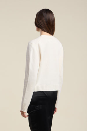 A person with long brown hair is seen from behind, standing against a plain beige background, wearing the ETNA CARDIGAN by Velvet by Graham & Spencer—a white wool blend cardigan with a V-neckline—paired with black pants.