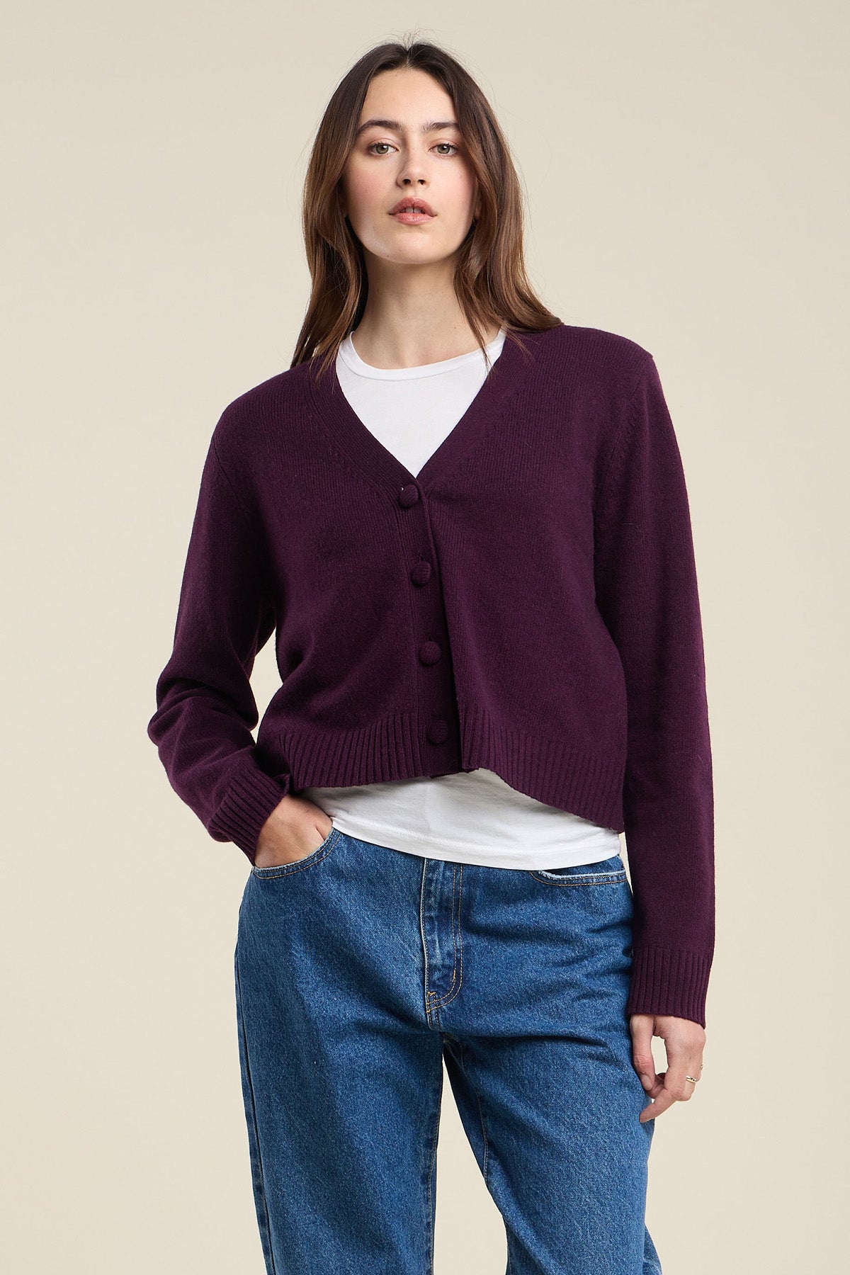 A woman stands against a plain beige background wearing the ETNA CARDIGAN by Velvet by Graham & Spencer—a dark purple wool blend cardigan with a V-neckline and tailored fit—layered over a white shirt and blue jeans.