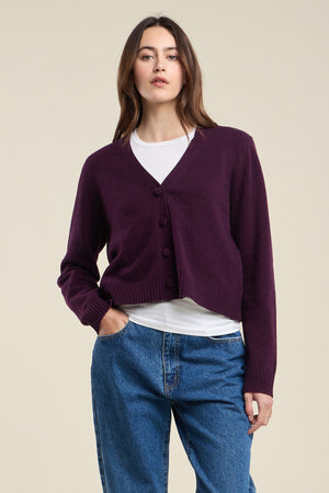 A woman stands against a plain beige background wearing the ETNA CARDIGAN by Velvet by Graham & Spencer—a dark purple wool blend cardigan with a V-neckline and tailored fit—layered over a white shirt and blue jeans.