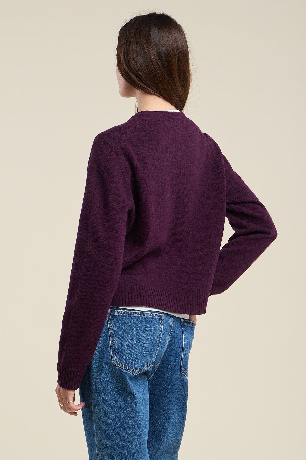 A person with long brown hair wears the ETNA CARDIGAN by Velvet by Graham & Spencer—a purple wool blend V-neck cardigan—paired with blue jeans, standing with their back to the camera against a plain background.-42063738470593