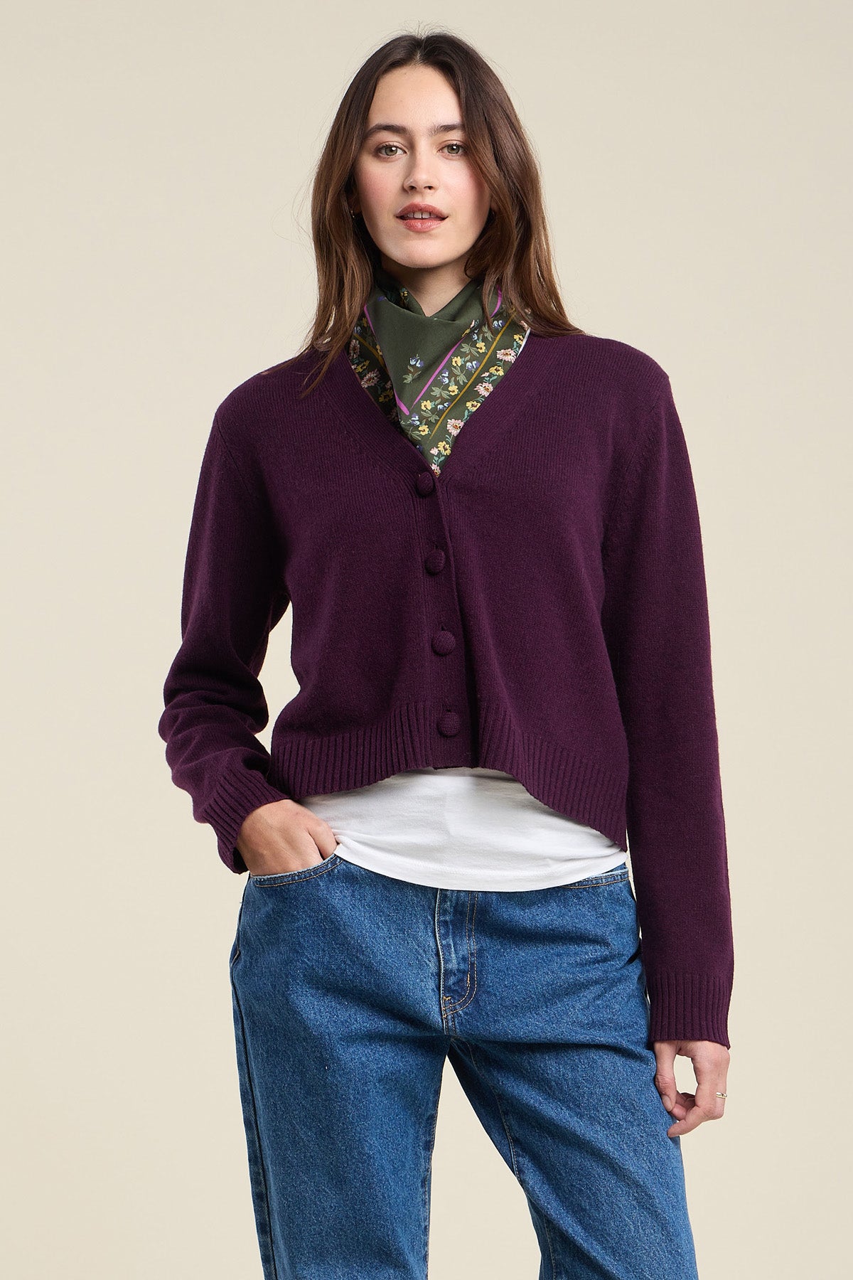 A woman poses against a plain background wearing the Velvet by Graham & Spencer ETNA CARDIGAN—a purple, tailored-fit wool blend cardigan with a V-neckline—layered over a white top, blue jeans, and finished with a green patterned scarf around her neck.