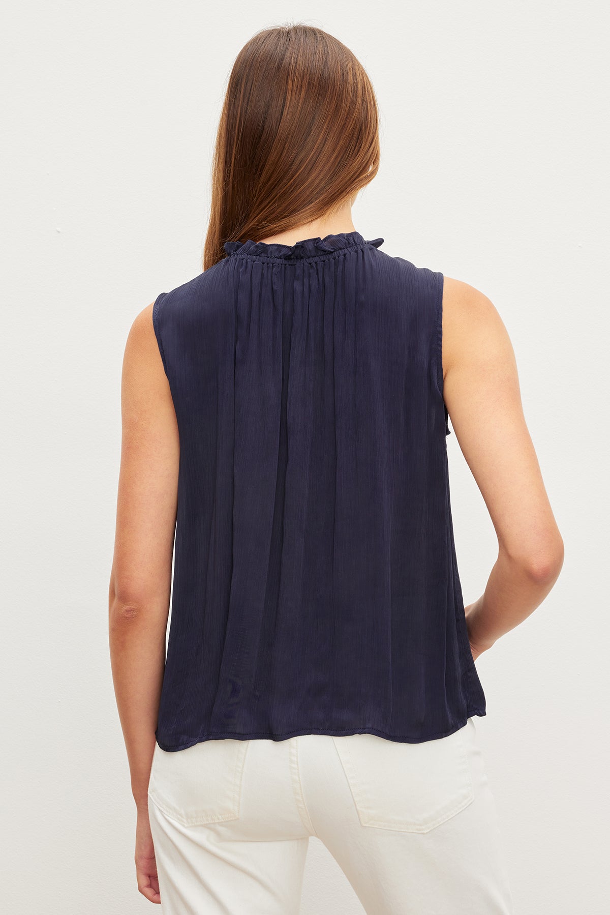   Woman seen from behind, wearing a navy KIANA RUFFLE NECK TANK TOP by Velvet by Graham & Spencer and white pants, standing against a neutral background. 