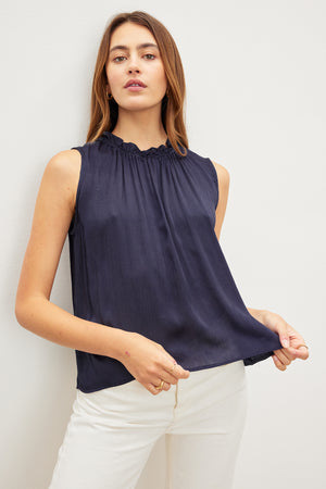 Woman in a navy blue KIANA RUFFLE NECK TANK TOP by Velvet by Graham & Spencer and white pants posing against a light background.