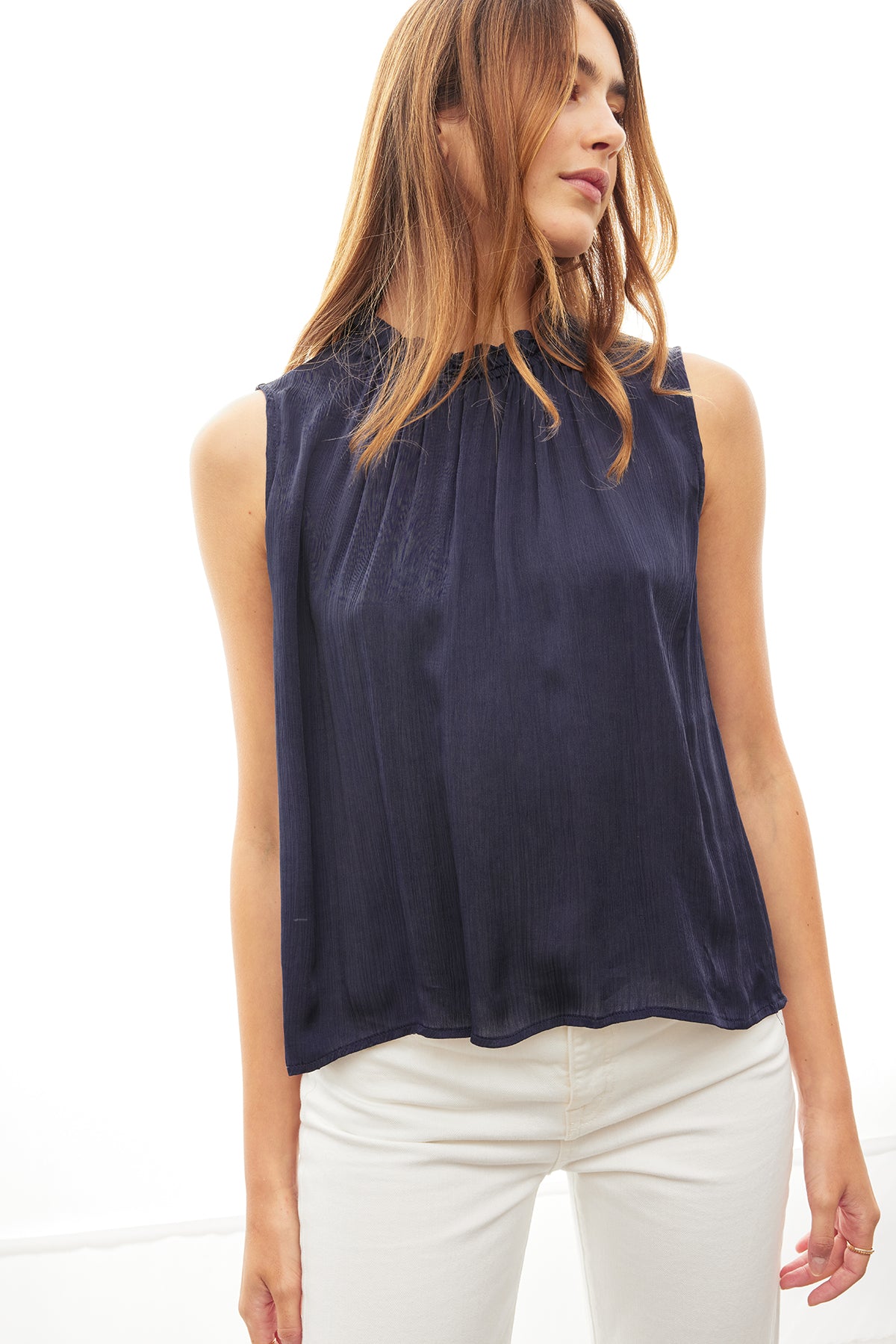   Woman in a KIANA RUFFLE NECK TANK TOP by Velvet by Graham & Spencer, navy blue tank top with an elastic ruffle neckline and white pants, standing against a white background. 