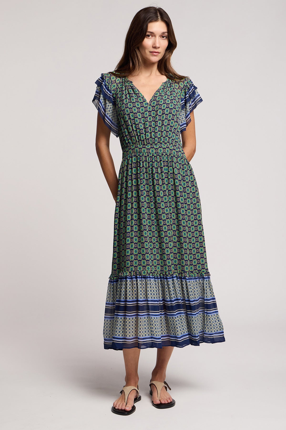 A woman wearing the CHERYL DRESS by Velvet by Graham & Spencer, a green and blue bold border print midi dress with short ruffled sleeves, paired with black sandals, stands against a plain background.-41526963044545
