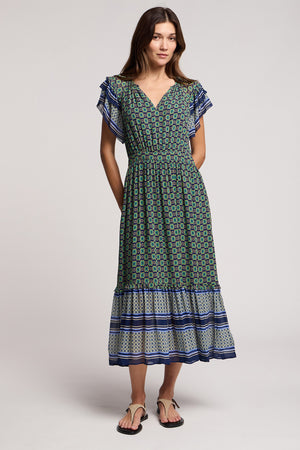 A woman wearing the CHERYL DRESS by Velvet by Graham & Spencer, a green and blue bold border print midi dress with short ruffled sleeves, paired with black sandals, stands against a plain background.