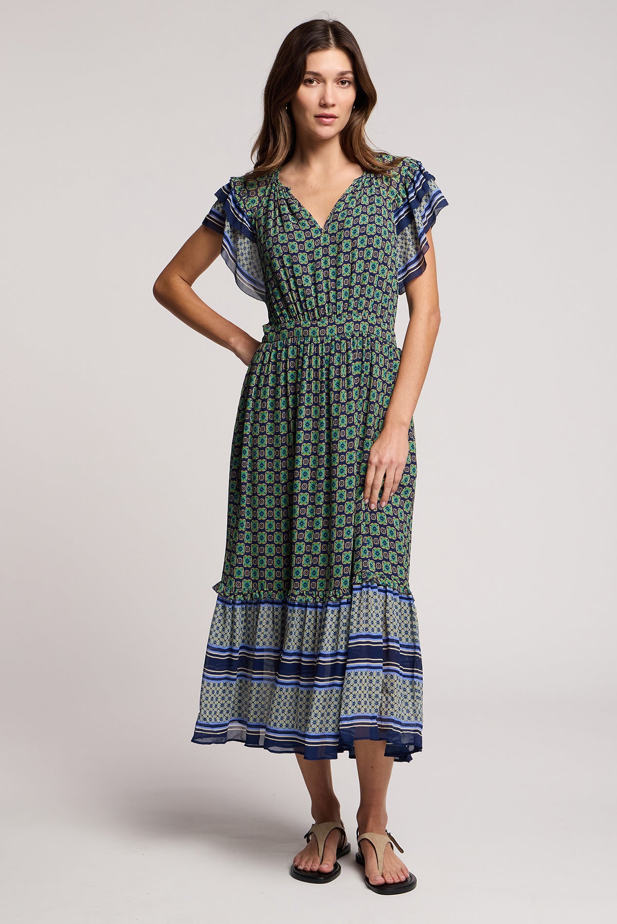 A woman wears the CHERYL DRESS by Velvet by Graham & Spencer, a resort-ready green and blue patterned midi dress with flutter sleeves and a bold border print at the striped hem, paired with black sandals.