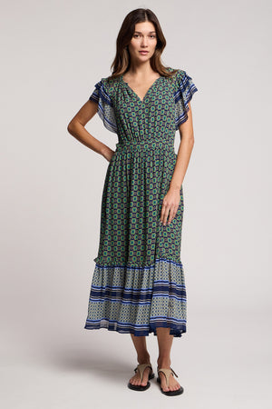 A woman wears the CHERYL DRESS by Velvet by Graham & Spencer, a resort-ready green and blue patterned midi dress with flutter sleeves and a bold border print at the striped hem, paired with black sandals.