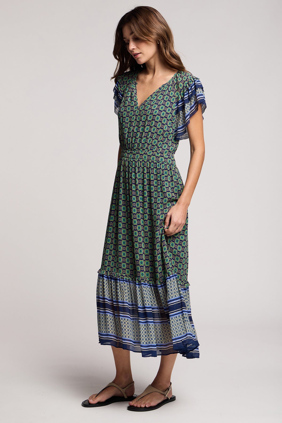 A woman models the Velvet by Graham & Spencer CHERYL DRESS, a resort-ready midi with short flutter sleeves, V-neckline, and bold border print. She completes the look with sandals against a plain background.