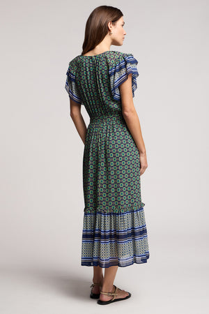 A woman stands with her back turned, wearing the CHERYL DRESS by Velvet by Graham & Spencer—a resort-ready midi style featuring short ruffled sleeves and a striking blue and white border-print hem, paired with flat sandals.