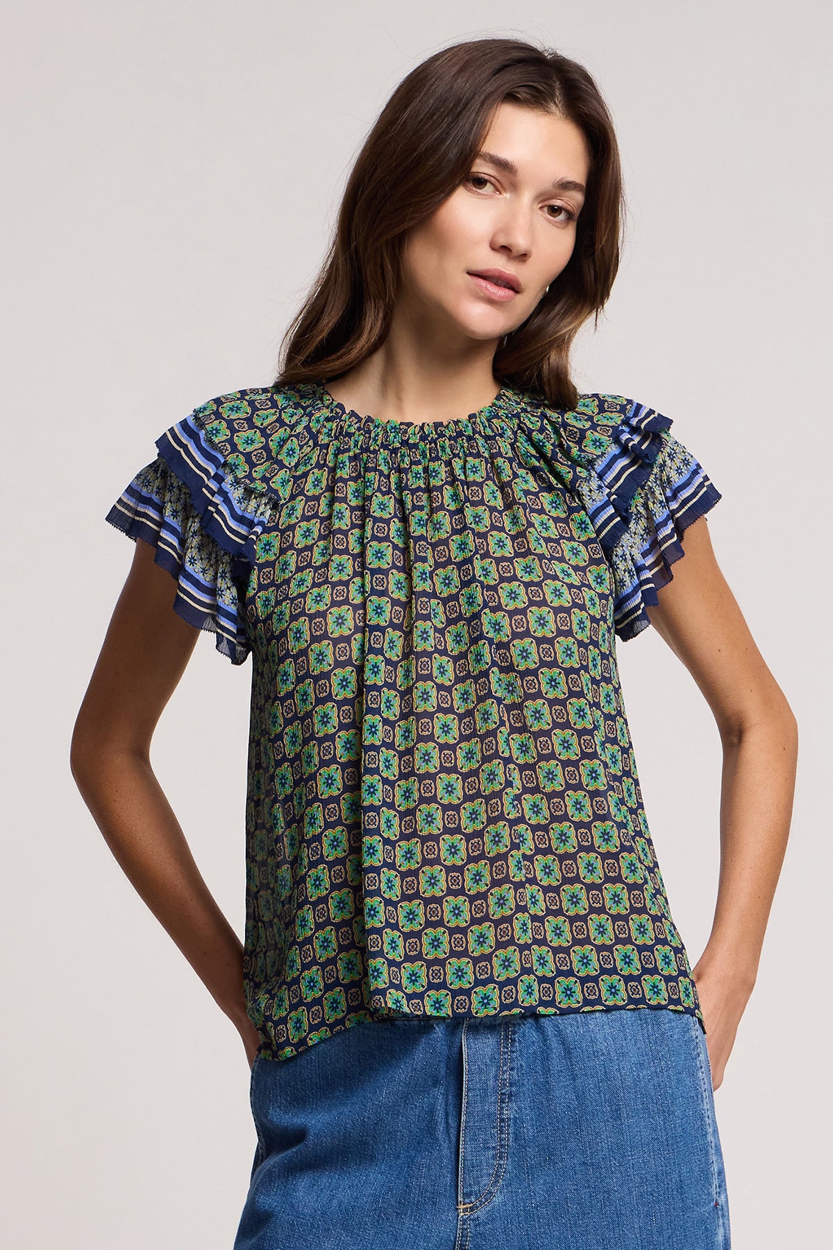 A woman wearing the DAHLIA TOP by Velvet by Graham & Spencer—a patterned, crinkled viscose short-sleeve blouse with ruffled shoulders—and blue denim jeans stands facing the camera against a plain background.-41526968844481