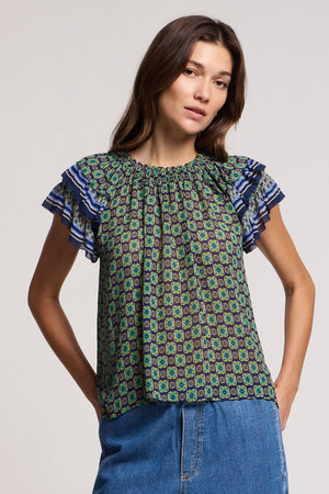 A woman wearing the DAHLIA TOP by Velvet by Graham & Spencer—a patterned, crinkled viscose short-sleeve blouse with ruffled shoulders—and blue denim jeans stands facing the camera against a plain background.