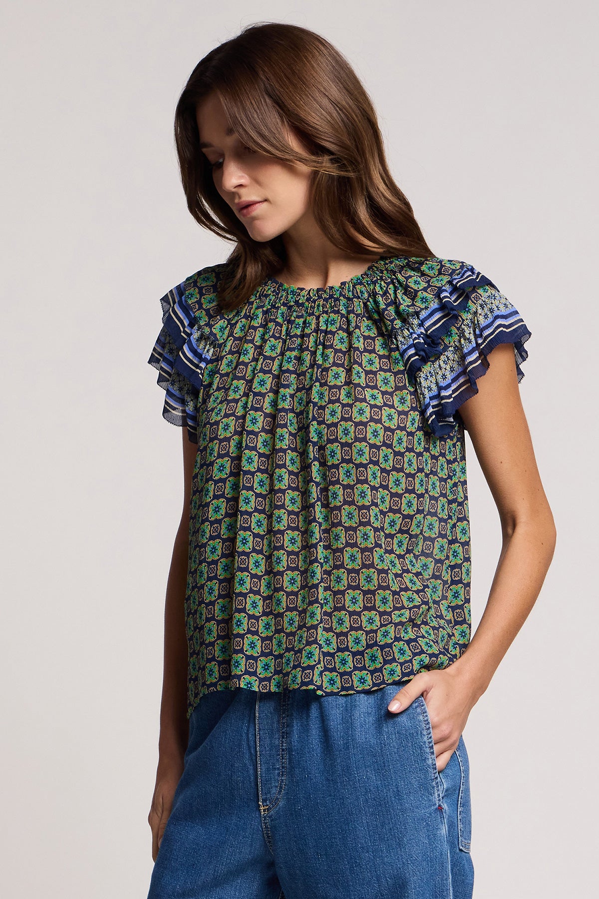 A woman wears the DAHLIA TOP by Velvet by Graham & Spencer, a green and blue patterned crinkled viscose blouse with ruffled shoulders and border print, paired with blue jeans, standing with one hand in her pocket.