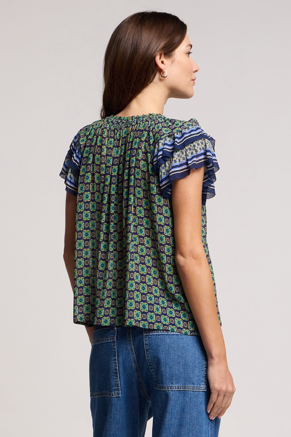 A woman faces away in the DAHLIA TOP by Velvet by Graham & Spencer—a patterned crinkled viscose blouse with ruffled short sleeves—paired with blue jeans, set against a neutral background.-41526968910017