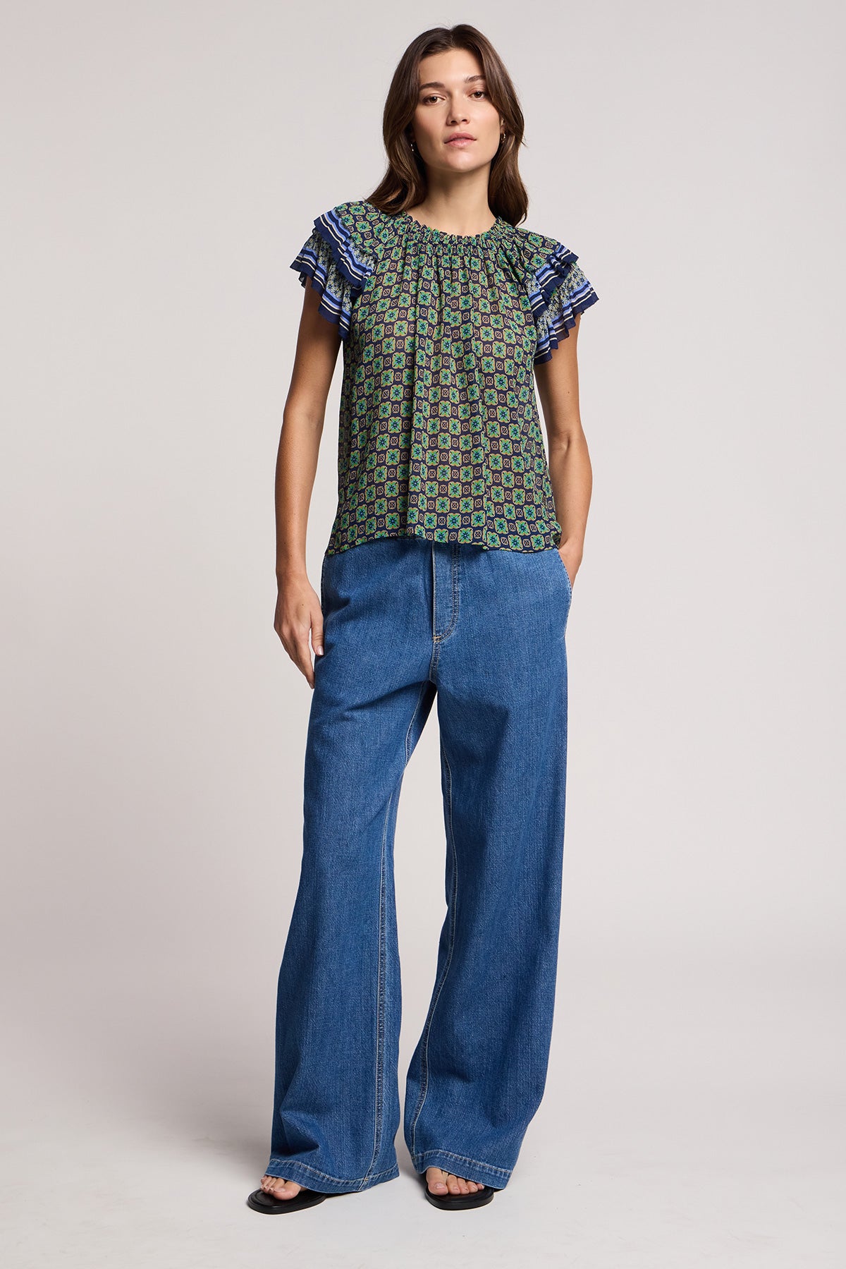 A woman stands in a studio wearing the DAHLIA TOP by Velvet by Graham & Spencer, a patterned crinkled viscose short-sleeve blouse with ruffled sleeves, paired with wide-leg blue jeans and black open-toe shoes.
