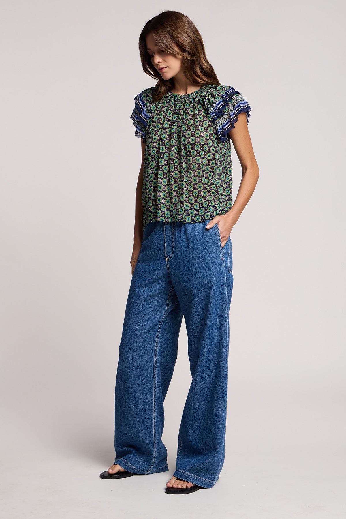 A woman stands in a relaxed pose against a plain light background, wearing a patterned blouse with ruffled sleeves, black sandals, and the SYD JEAN by Velvet by Graham & Spencer—wide-leg denim jeans featuring an elastic drawstring waist for a comfortable, relaxed fit.