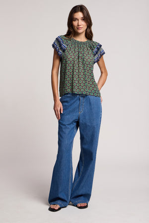A woman stands facing forward against a plain background, wearing a patterned short-sleeve blouse with ruffled sleeves, the SYD JEAN by Velvet by Graham & Spencer—relaxed fit wide-leg denim jeans featuring an elastic drawstring waist—and open-toe black sandals.