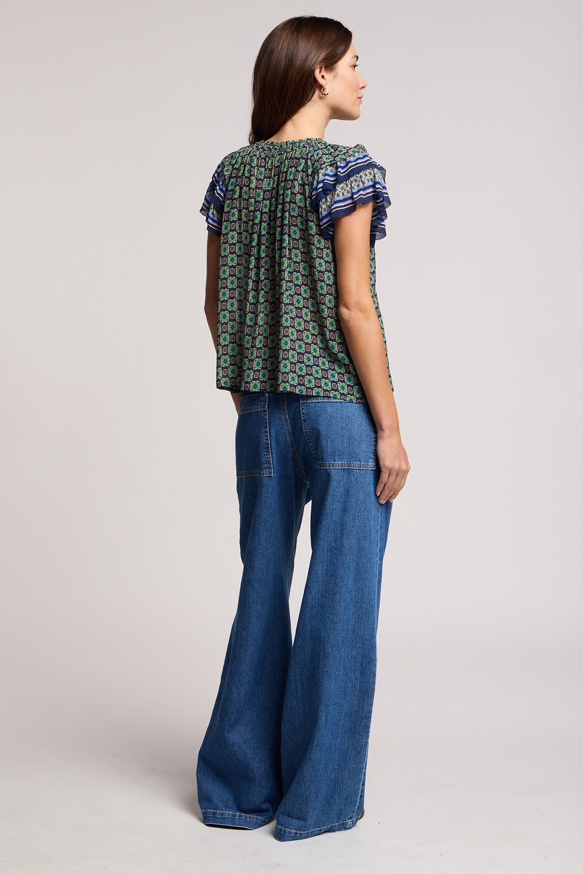 A woman stands facing away against a plain, light background, wearing the Velvet by Graham & Spencer SYD JEAN—a relaxed-fit, wide-leg denim style with an elastic drawstring waist—paired with a patterned short-sleeve top.