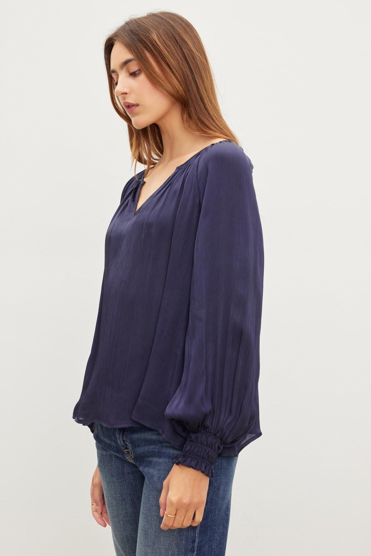 A woman with long hair wears a loose-fitting, long-sleeved dark blue HELEN TOP by Velvet by Graham & Spencer and blue jeans, standing against a plain white background.-37073685053633